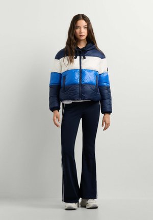 Junge Frau in blau-weiß-navyfarbiger Daunenjacke, schwarzen ausgestellten Hosen mit weißen Streifen und weißen Sneakers steht vor einem einfarbigen Hintergrund.