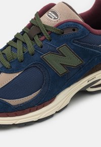 Navy suede and mesh sneaker featuring a green logo, brown accents, and textured laces. Cushioned sole with a multicolored tread pattern.