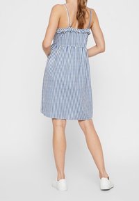 Robe rayée bleue et blanche avec fines bretelles et encolure à volants ; coupe décontractée, longueur genou, fabriquée en tissu léger. Chaussures blanches.