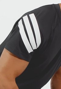 Camiseta atlética negra con rayas blancas perforadas en los hombros. El tejido parece ligero y tiene una textura suave.