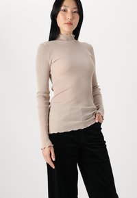 Haut beige à manches longues à côtes, avec un col haut à volants et un ourlet festonné, associé à un pantalon en velours côtelé noir. Texture lisse, design ajusté.