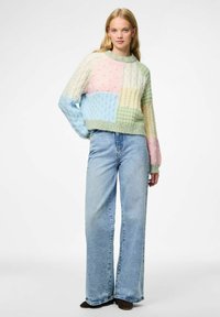 Sweter dzianinowy z pastelowym patchworkiem w odcieniach różu, niebieskiego, zielonego i kremowego; teksturowany wzór w warkocze; w połączeniu z jasnoniebieskimi, szerokimi spodniami.