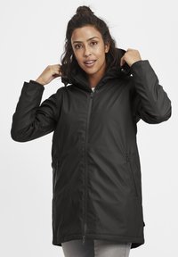 Chaqueta negra impermeable con capucha, frontal con cremallera, dos bolsillos laterales y puños elásticos. Textura suave con un acabado ligeramente brillante.