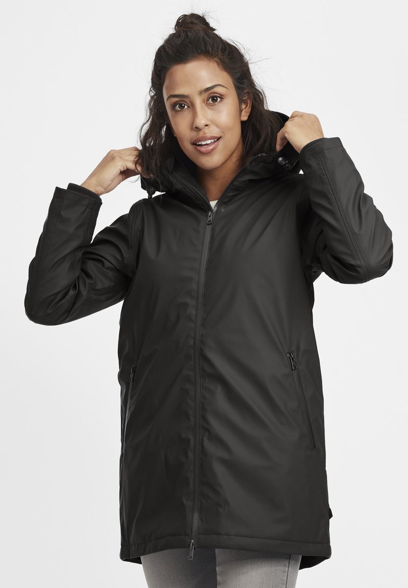 Chaqueta negra impermeable con capucha, frontal con cremallera, dos bolsillos laterales y puños elásticos. Textura suave con un acabado ligeramente brillante.