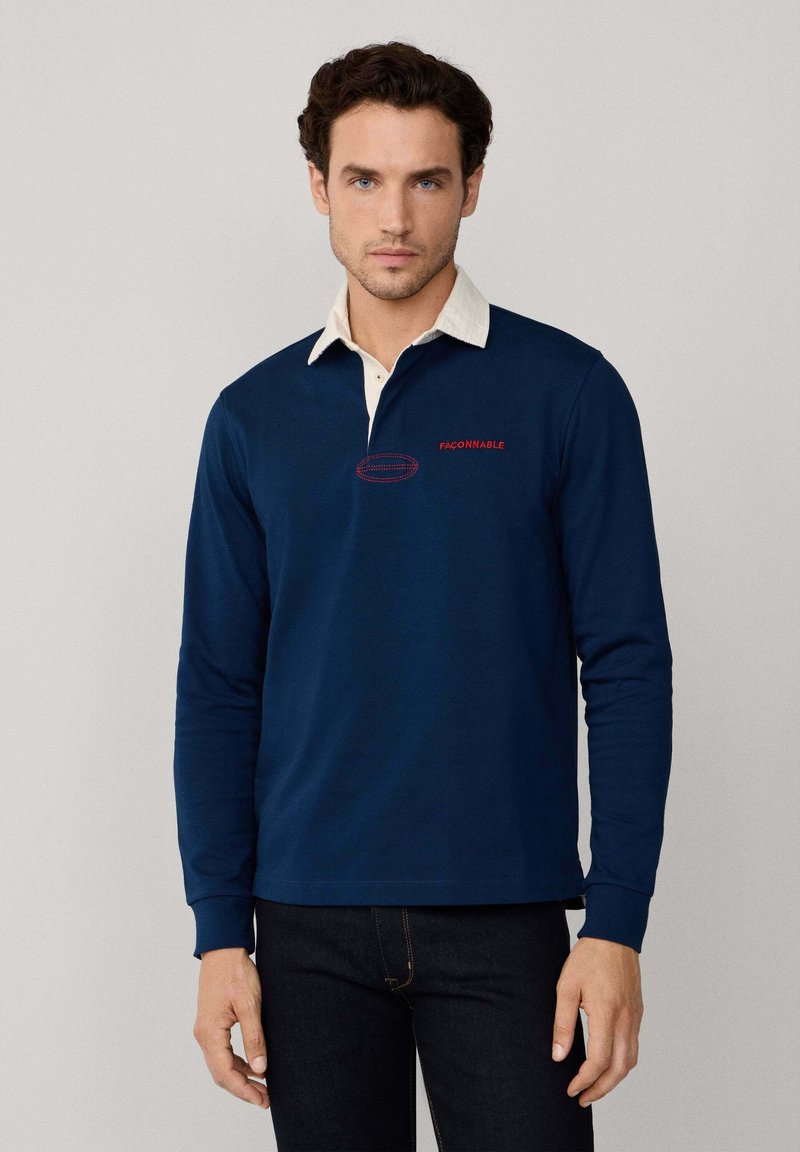 Mann in navyblauem Langarm-Polohemd mit weißem Kragen und rotem "Façonnable"-Text, kombiniert mit dunklen Jeans, steht vor einem einfarbigen Hintergrund.