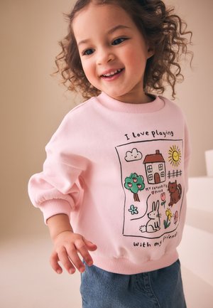 Usmievajúce sa batoľa oblečené v ružovej mikine s hravými obrázkami zvierat a prírody a textom, v kombinácii s modrými džínsami.