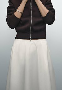 Felpa zip-up marrone scuro con polsini a coste abbinata a una gonna bianca plissettata, che mette in evidenza un tessuto liscio e una silhouette pulita.