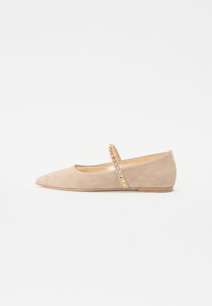 Chaussure plate en daim beige avec un bout pointu, dotée d'une sangle décorative ornée de clous dorés pour l'accent.