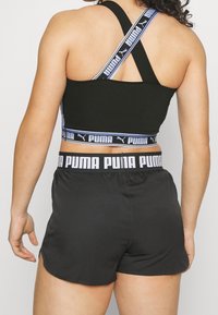 Schwarzer Sport-BH mit Cross-Back-Design; verfügt über ein blau-schwarzes Markenband. Kombiniert mit schwarzen Sportshorts. Glatte, eng anliegende Textur.