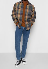 Rutigt flanellskjorta i blått, beige och grönt, med två bröstfickor, bärs över en brun tröja, ihop med blå jeans och svarta sneakers.