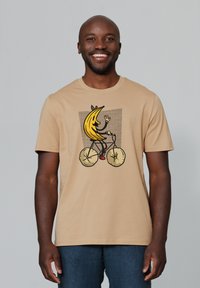 Beige t-shirt met een afbeelding van een cartoonachtige banaan die op een fiets rijdt, met een gestructureerde achtergrond en kleurrijke details.