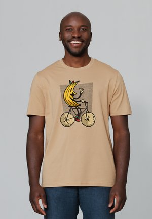 T-shirt beige avec un graphisme représentant une banane cartoon faisant du vélo, sur un fond texturé et avec des détails colorés.