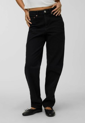 Persoon draagt losse zwarte jeans met hoge taille, handen op heupen, zwarte ballerina's op een effen lichte achtergrond.