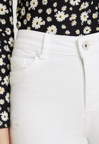 Jeans in denim bianco con vita alta, dotati di una tasca frontale, accessori in argento e una toppa strappata sulla gamba. Top floreale nero visibile sopra.