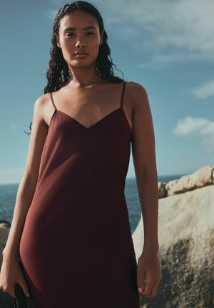 Žena s dlhými tmavými kučeravými vlasmi, oblečená v bezrukávové bordovej šaty, stojaca pri kamenistej pláži s oceánom a zamračenou oblohou v pozadí.