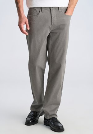 Mand iført teksturerede grå bukser med lige ben og sorte læderloafer, den ene hånd i lommen, stående på en ensartet lys baggrund.