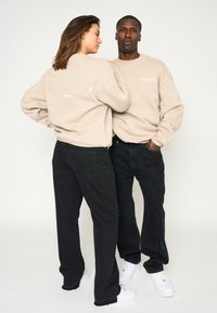 Zwei Personen in beigen Sweatshirts mit "Multiply"-Branding, schwarzen Jeans und weißen Sneakern, die eng zusammen vor einem weißen Hintergrund posieren.