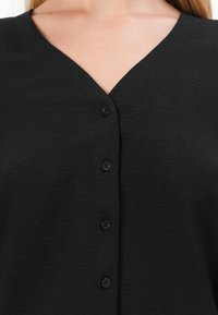 Svart knapp-up topp med V-hals, med teksturert stoff og fem svarte knapper foran. Enkel og udetaljert design.
