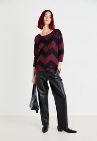 Maroon- und schwarze Chevron-musterlose Strickoberteil mit Dreiviertelärmeln, kombiniert mit schwarzen Lederhosen und spitz zulaufenden Stiefeln.