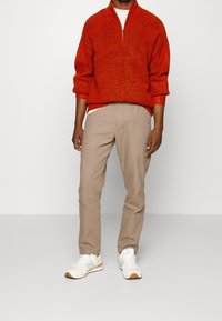 Roter Strickpullover mit Reißverschlusskragen, kombiniert mit beigen Chinos und weißen Sneakers mit orangefarbenen und blauen Akzenten. Sichtbare detaillierte Nähte.