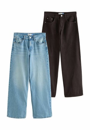 Žena oblečená v svetlomodrých voľných džínsoch s vysokým pásom a ružovom saténovom topu, s čipkovaným spodným prádlo viditeľným nad pásom.