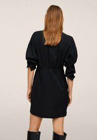Abito nero con vita stretta, maniche gonfie e taglio dritto. Realizzato in tessuto texture, presenta polsini con bottoni e un design minimalista.