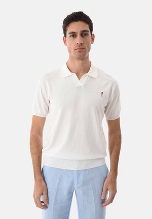 Giovane uomo di fronte, indossa una polo bianca a maniche corte e pantaloni azzurri chiari su uno sfondo chiaro semplice.