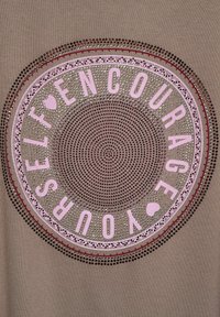 Brauner Stoff mit einem kreisförmigen Grafikdesign, auf dem der Text "SELBST ERMUtIGEN SIE IHR" in Pink steht, umgeben von gestickter Spitze und Strasssteinen.