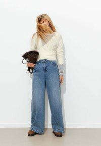 Pull en tricot blanc par-dessus une chemise rayée, associé à un jean bleu à jambe large et des chaussures à imprimé léopard. Le mannequin tient un sac marron.