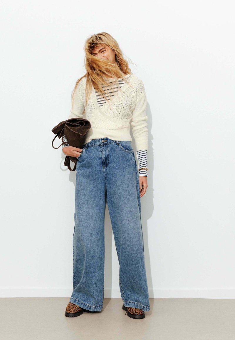 Pull en tricot blanc par-dessus une chemise rayée, associé à un jean bleu à jambe large et des chaussures à imprimé léopard. Le mannequin tient un sac marron.