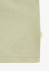 Tissu vert clair avec des coutures visibles à l'ourlet et une petite étiquette blanche avec un texte orange sur le bord droit.