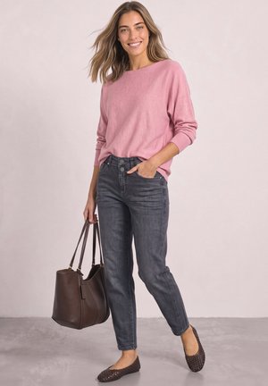 Kvinde smiler iført lyserød sweater, mørkegrå jeans, flade brune sko, holder stor brun håndtaske med den ene hånd i lommen.