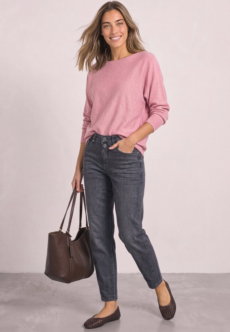 Femme souriante portant un pull rose, un jean gris foncé, des chaussures plates marron, tenant un grand sac à main marron avec une main dans la poche.