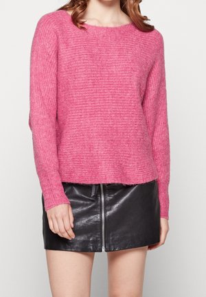 Femme portant un pull en maille rose à manches longues et une mini-jupe en cuir noire avec une fermeture éclair à l'avant, debout devant un fond uni.