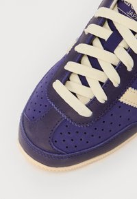 Baskets violettes avec une tige en cuir lisse, lacets crème, détails perforés et une semelle en caoutchouc beige. Présente un élément de design subtil.