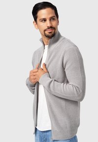 Cárdigan gris con relieve, con cremallera y cuello alto, mangas largas y bolsillos laterales, llevado sobre una camisa blanca. Presenta un diseño de tejido texturizado.