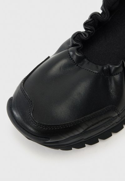 Black leather slip-on shoe with a ruched upper, rounded toe, and a textured sole for grip. Smooth finish with subtle stitching details.