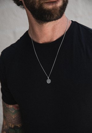 Sølvvedhæng halskæde på en tynd kæde, med en cirkulær charm, båret over en sort t-shirt. Synlige tatoveringer på armen.