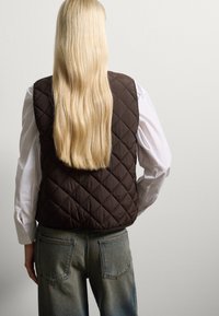 Gilet marrone trapuntato con colletto alto, indossato sopra una camicia bianca. Il gilet presenta un motivo a rombi e una texture liscia. Sono visibili jeans di denim blu.