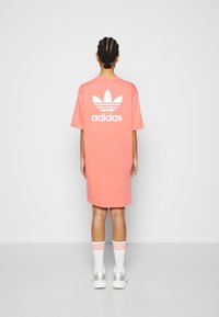 Průmyslově velké růžové bavlněné tričkové šaty s bílým logem Adidas a trojúhelníkovým znakem na zádech. Modelka má na nohou bílé ponožky s růžovými pruhy a tenisky.