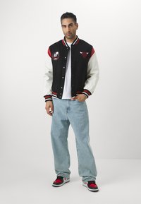 Melna un balta studentu jakas ar sarkaniem akcentiem, ar logotipiem; vilkta pāri baltai kreklam un gaišzilām džinsām, apģērbta ar sarkaniem un melniem sporta apaviem.