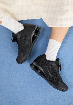 Ben iført hvide sokker og sorte Nike-sneakers, der hviler på et lyseblåt tæppe, med en hvid tekstureret nederdel synlig.