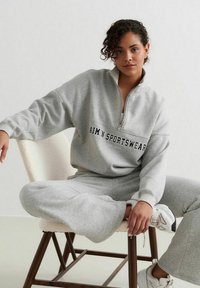 Gray zip-up sweatshirt with "AIM'N SPORTSWEAR" text on chest, paired with matching gray pants and white sneakers, seated on a wooden chair.