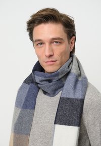 Uld tørklæde med et ternet mønster i nuancer af blå, grå og beige, med en blød tekstur og et loop-design omkring halsen.