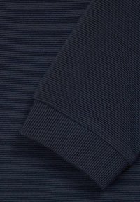 Detailní záběr na tmavě modrou ribbovanou pleteninu s ohnutým manžetovým detailem, který zvýrazňuje texturu a prošívání.