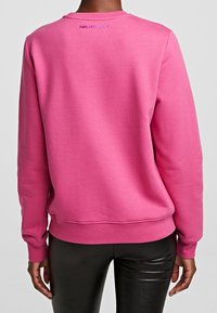 Sudadera rosa elaborada con una tela suave, con cuello redondo y puños acanalados. Logo bordado en morado cerca del cuello.