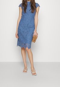 Niebieska koronkowa sukienka z krótkimi rękawami, falbaniastym dołem, skomplikowanym kwiatowym wzorem, w połączeniu z brązową teksturowaną kopertówką i białymi sandałami na cienkich paskach.
