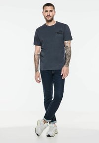 Tmavě šedé tričko s krátkým rukávem a logem na levé části hrudi, kombinované s tmavými džíny a bílými teniskami. Viditelné tetování na pažích.