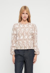 Jauna moteris su tiesiais šviesiais plaukais, vilkinti smėlio spalvos ilgomis rankovėmis palaidine su baltu gėlių išsiuvinėjimu ir tamsiomis plačiomis kelnėmis.