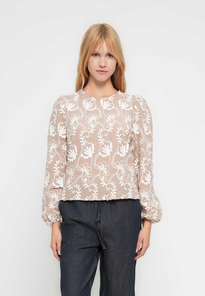 Jauna moteris su tiesiais šviesiais plaukais, vilkinti smėlio spalvos ilgomis rankovėmis palaidine su baltu gėlių išsiuvinėjimu ir tamsiomis plačiomis kelnėmis.